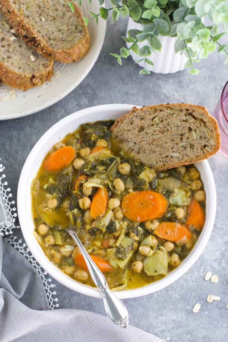 A white bowl filled with soup and a slice of bread next to a plant on a gray background. 