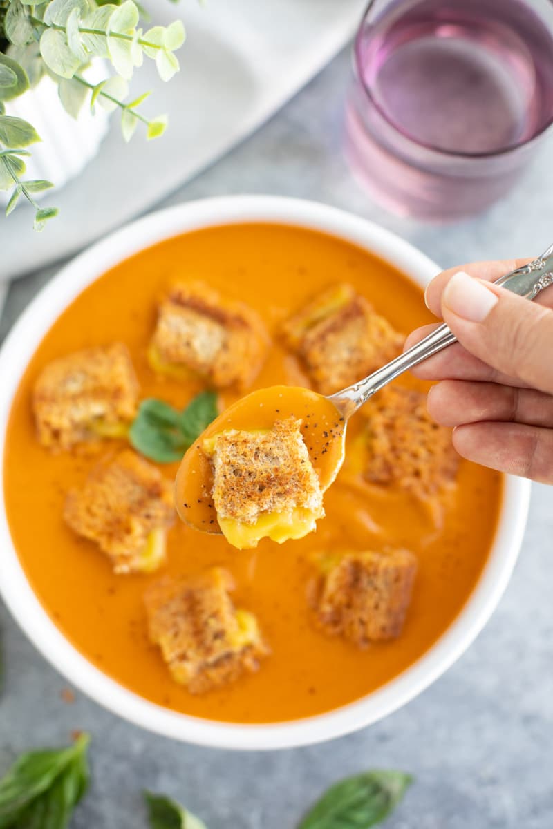 A hand holding a spoonful of soup and a grilled cheese crouton on a gray background.