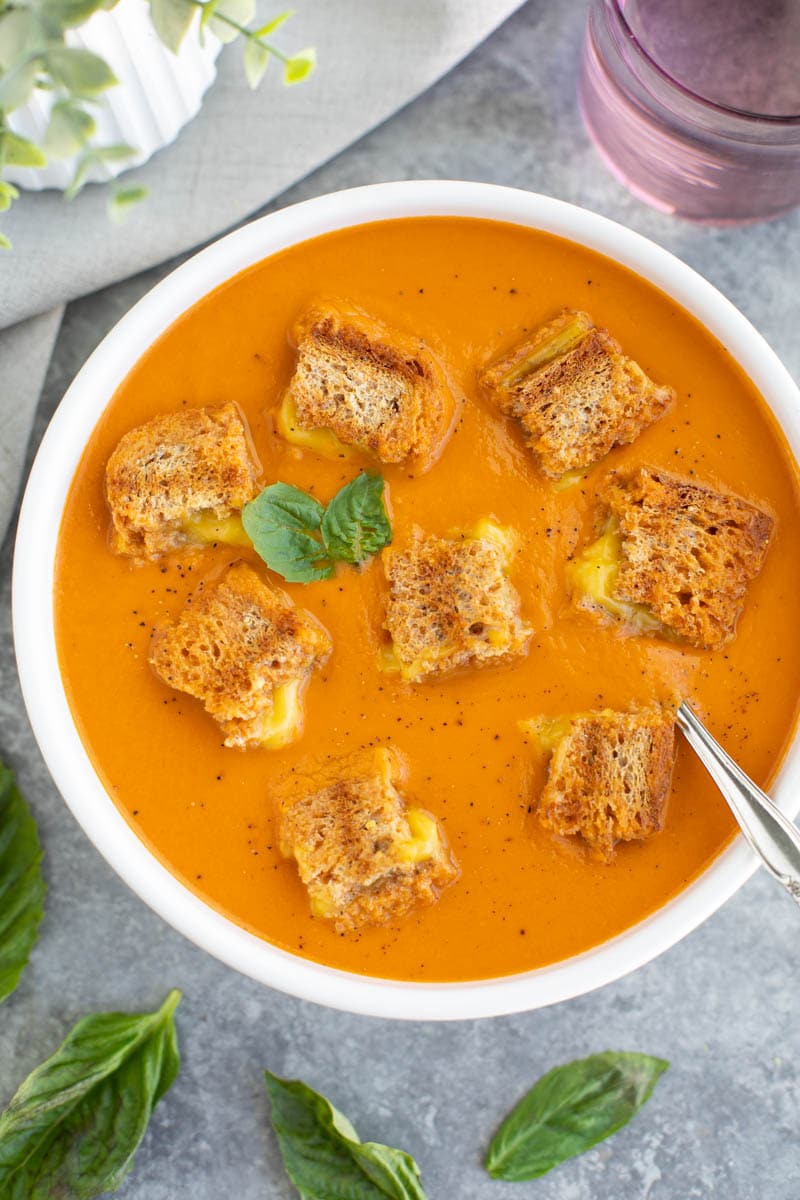 A white bowl filled with tomato soup and a spoon and it's topped with grilled cheese croutons on a gray background.