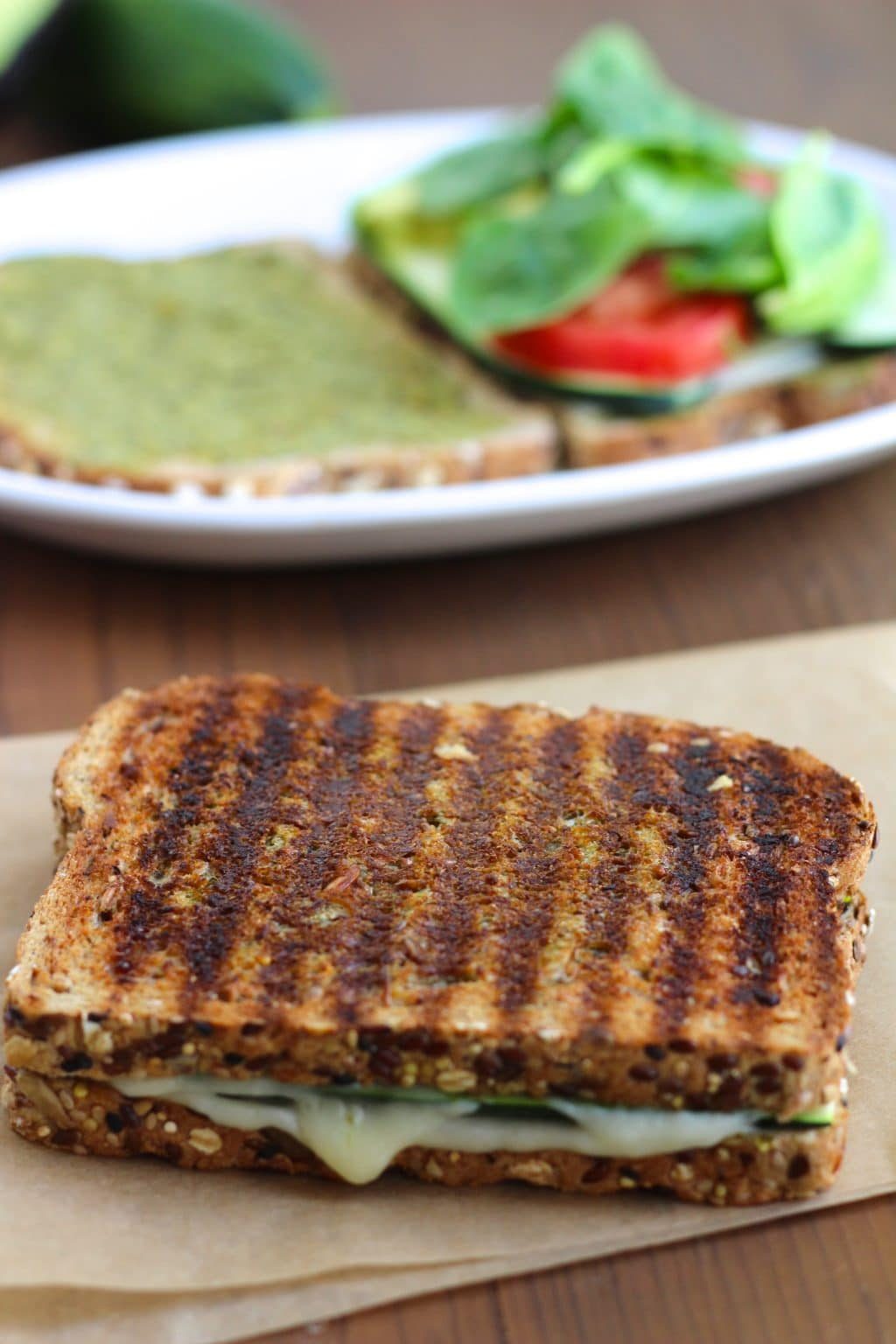 A pressed grilled cheese with dark grill marks on top of a piece of parchment paper with a white plate behind it on a rustic background. 