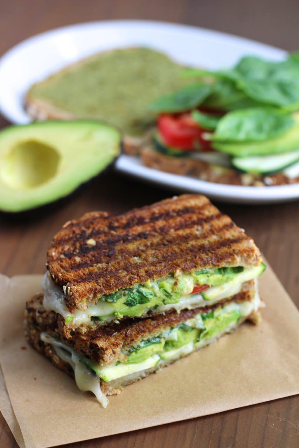 A sandwich cut in half and stacked on top of each other on top of a piece of parchment paper next to a white plate and half an avocado. 