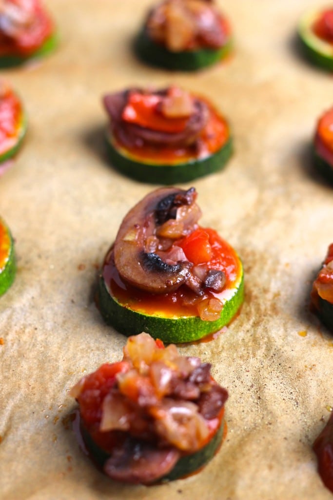 Slices of zucchini topped with tomato sauce and mushrooms on a parchment-lined tray.