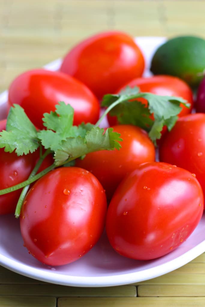 A bunch of wet Roma tomatoes, cilantro, and limes on a white plate with a wooden background.