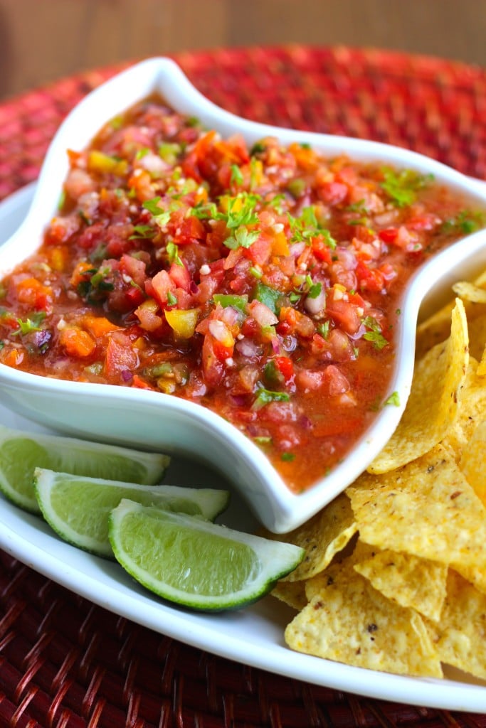 A white bowl filled with salsa next to chips and lime wedges on a white plate.