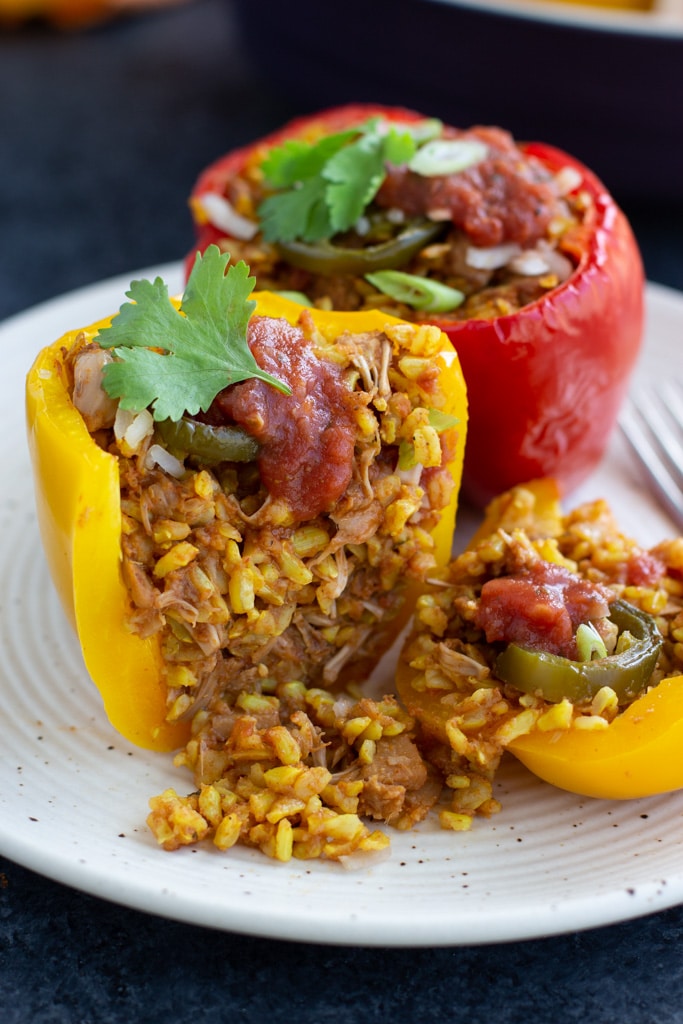 A yellow vegan stuffed bell pepper that is cut in half and the filling is falling out. There is also a full red stuffed pepper behind it sitting on a white textured plate.