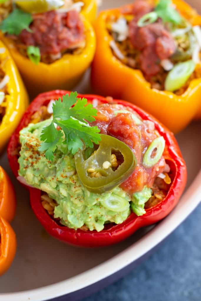 A close up shot of a vegan stuffed pepper in a casserole dish that's topped with guacamole, salsa, a jalapeño, and cilantro. 