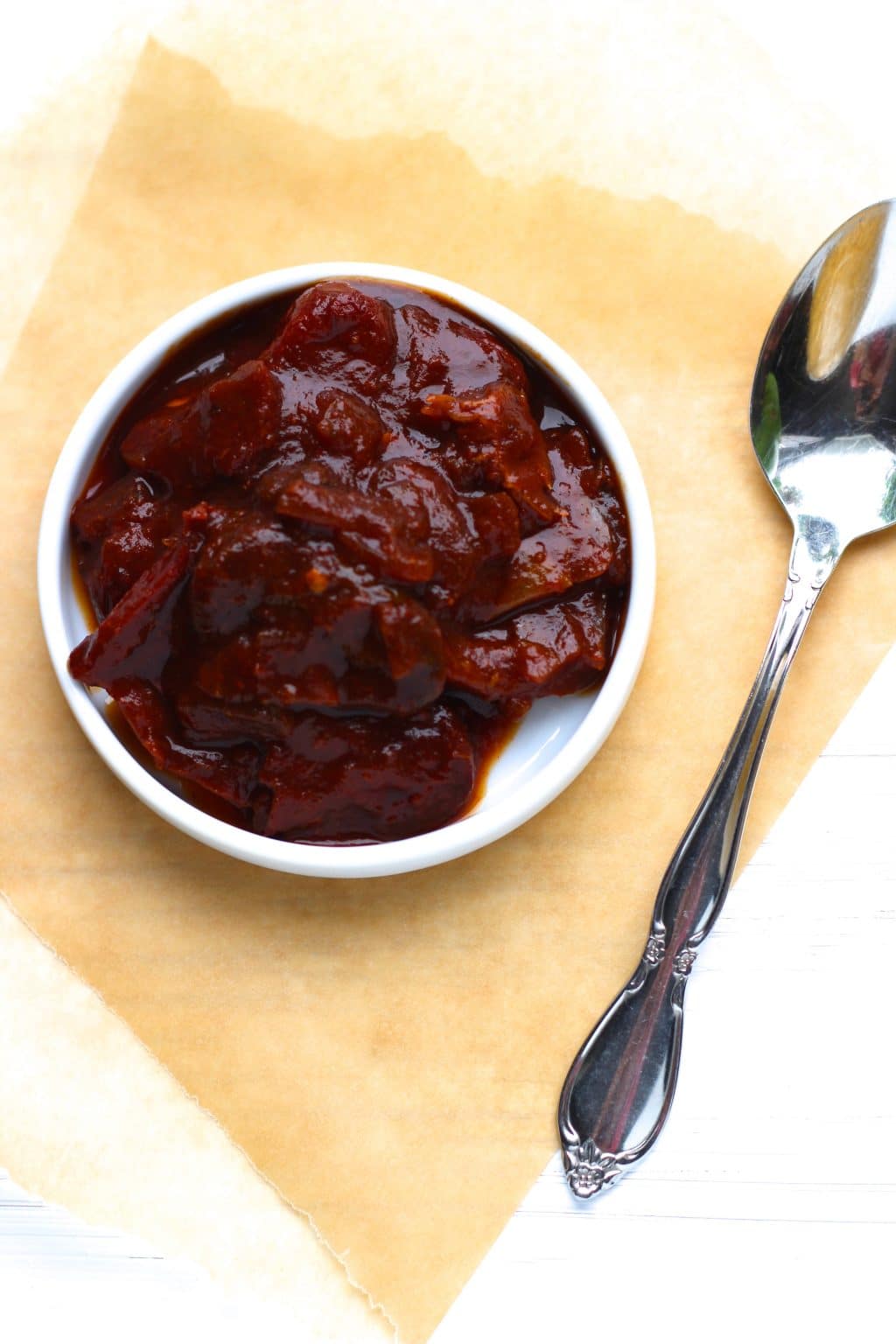 A small white dish filled with chipotle peppers next to a spoon on top of a piece of parchment paper.