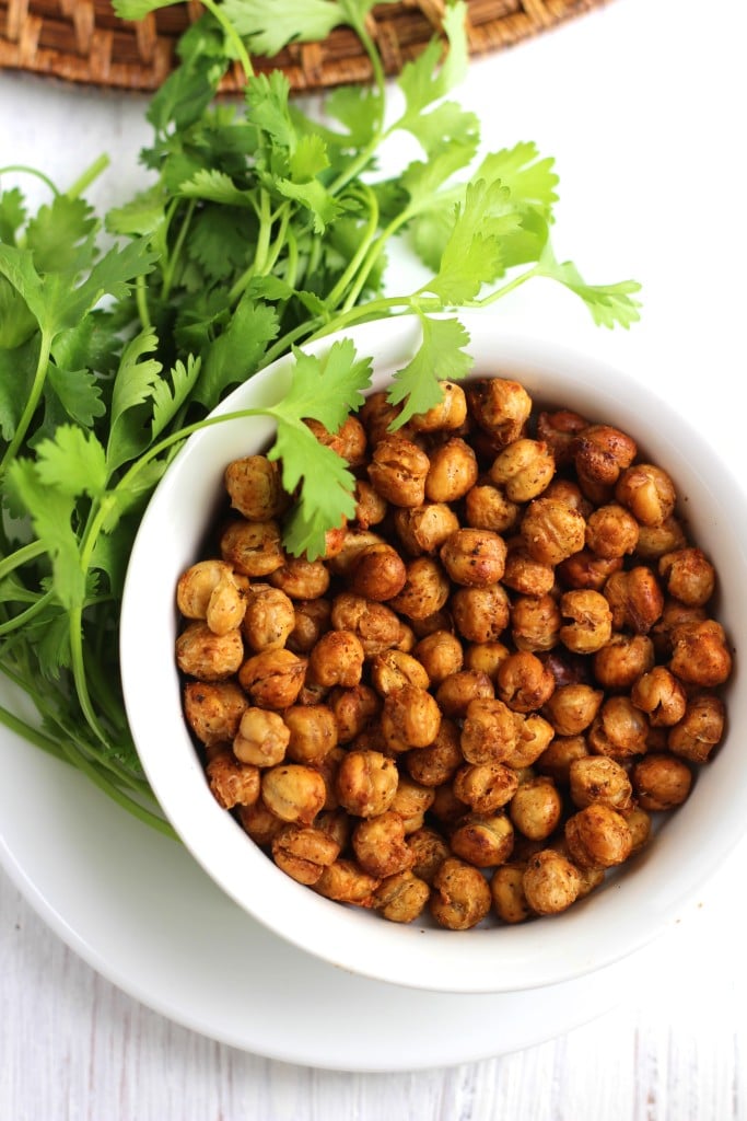A white bowl filled with roasted chickpeas next to a bunch of cilantro on a white background.