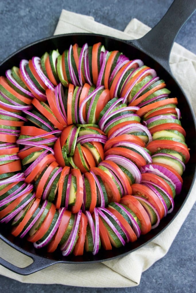 Overhead shot of thinly sliced vegetables in a circular pattern in a cast iron skillet. 