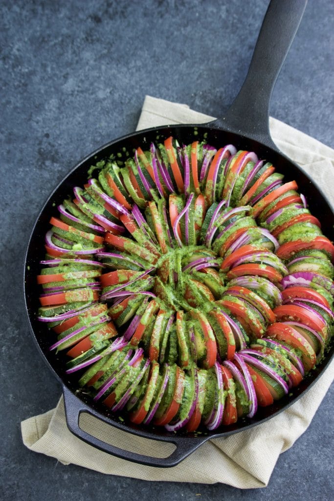 Overhead shot of sliced vegetables topped with pesto in a cast iron pan on a dark background.