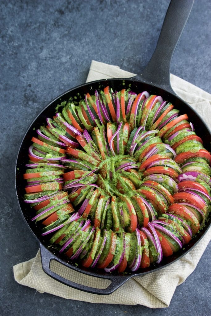 Overhead shot of sliced vegetables topped with pesto in a cast iron pan on a dark background.