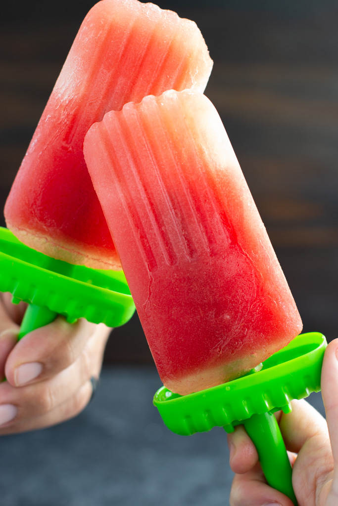 Two hands holding two homemade watermelon popsicles with green bases. 