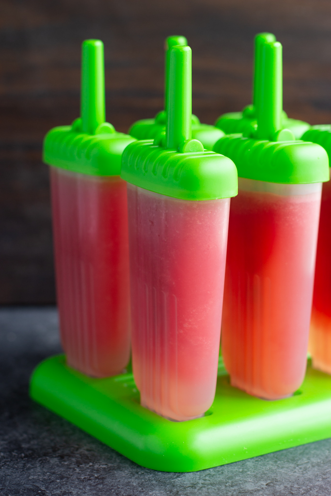 A tray of homemade watermelon popsicles on a green base resting on top of a textured table. 