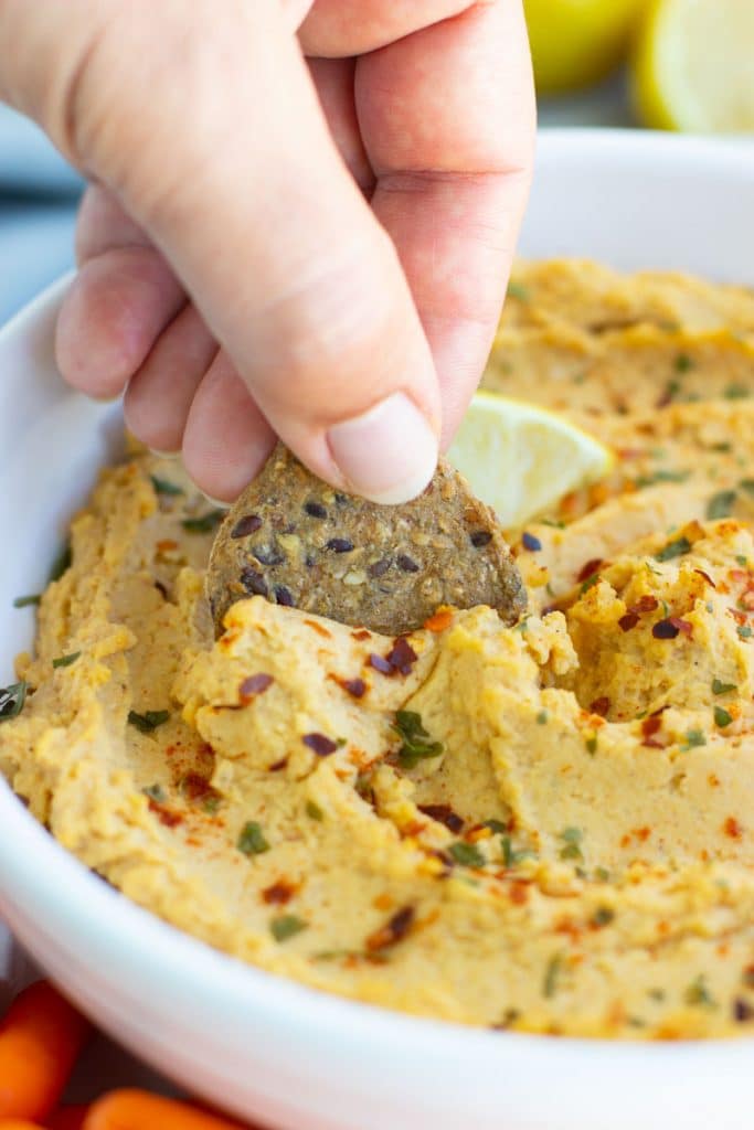 A hand dipping a cracker in a white bowl filled with hummus. 