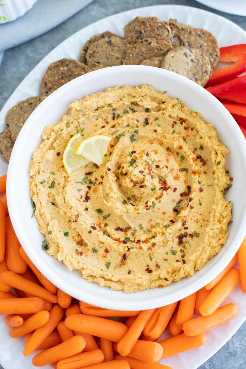 A white bowl filled with acorn squash hummus on a white platter with carrots, peppers, and crackers. 
