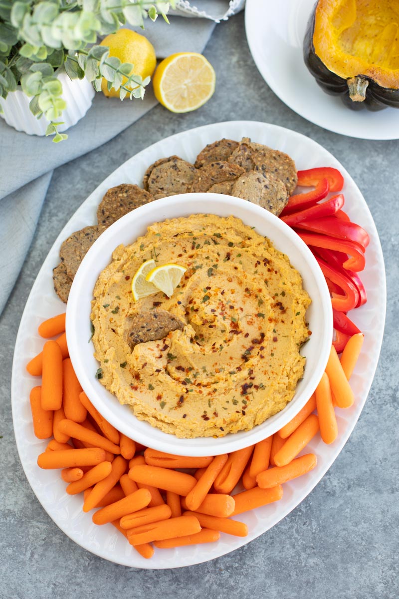 A white bowl filled with hummus on a white platter filled with crackers and veggies on a gray background.