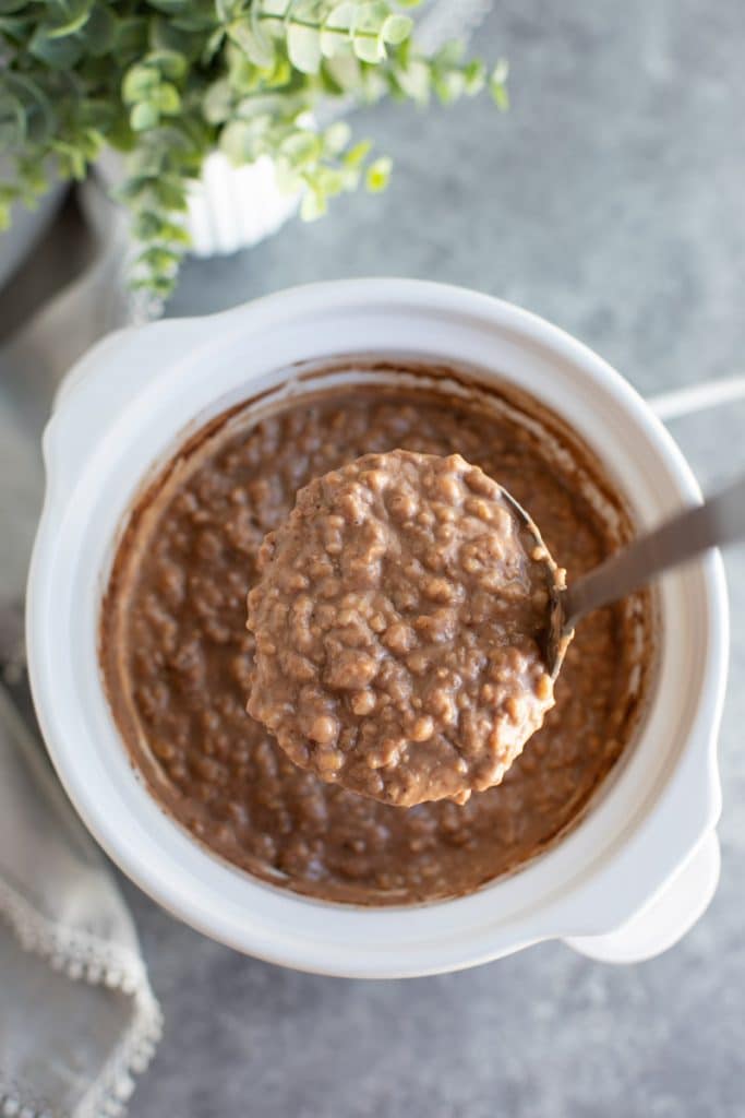 A white bowl filled with oatmeal, loose steel cut oats, teaspoons, and a protein shake on top of a wooden cutting board.