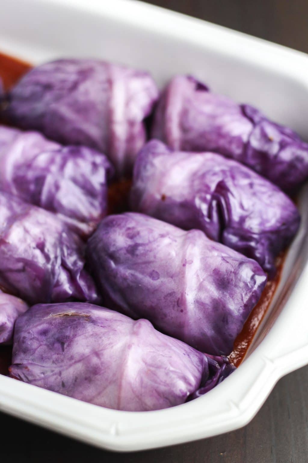 Stuffed red cabbage rolls sitting in tomato sauce inside a white casserole dish. 