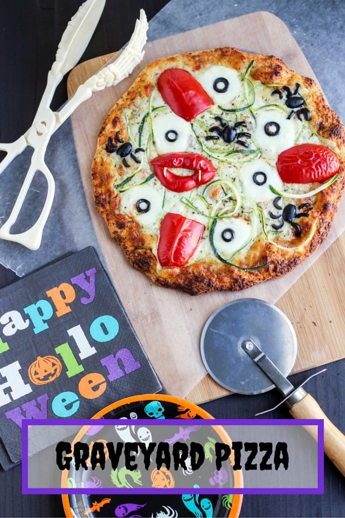 A pizza on a cutting board next to a pizza cutter, halloween napkin, and skeleton tongs. 