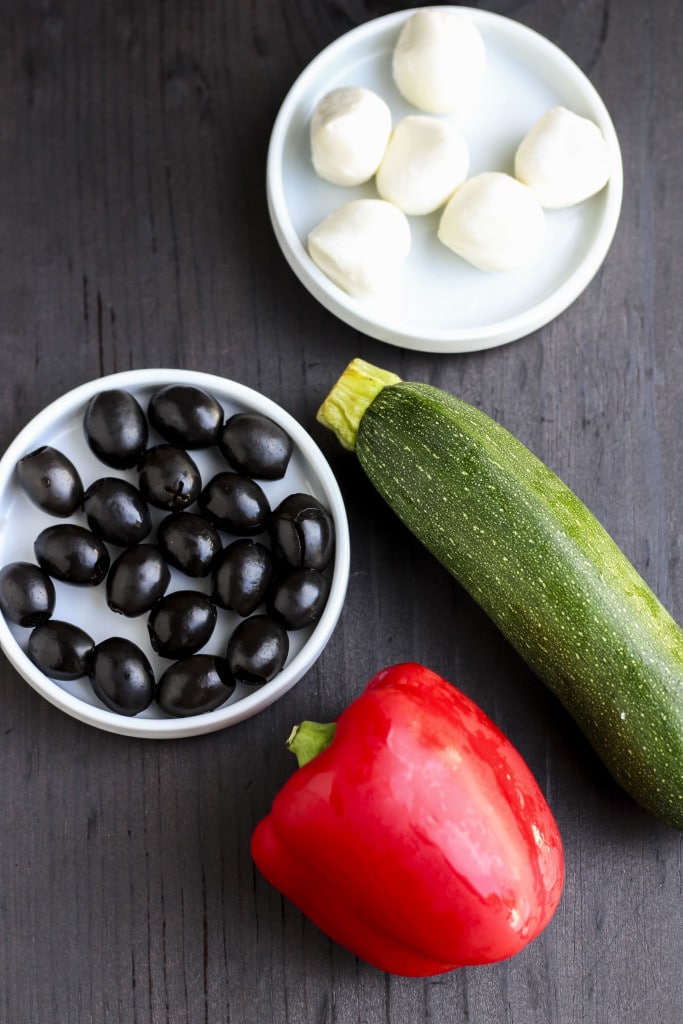 Mozzarella balls, olives, zucchini, and a red pepper on a dark background. 