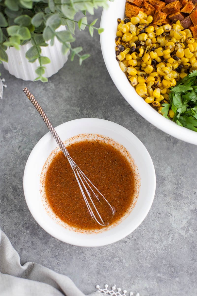 A small white bowl of dressing next to a plant and a bowl of vegetables on a gray background.