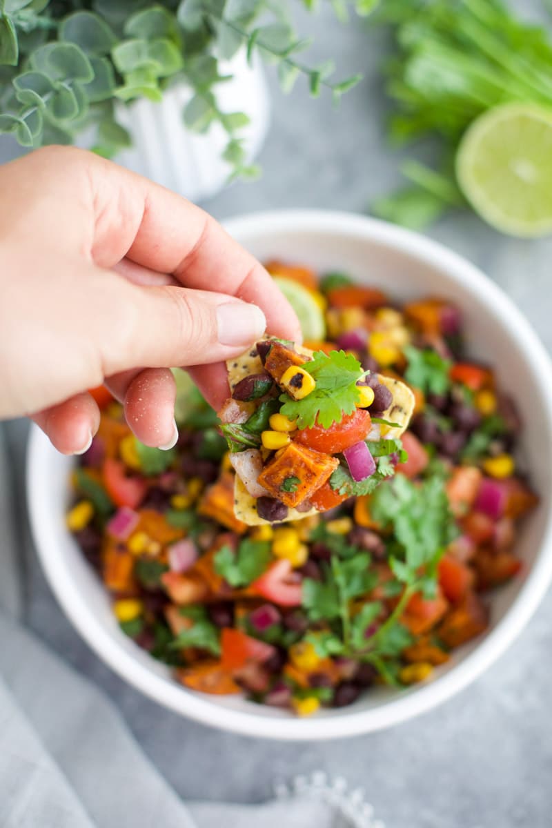 A hand dipping a chip into salsa over a white bowl on a gray background.