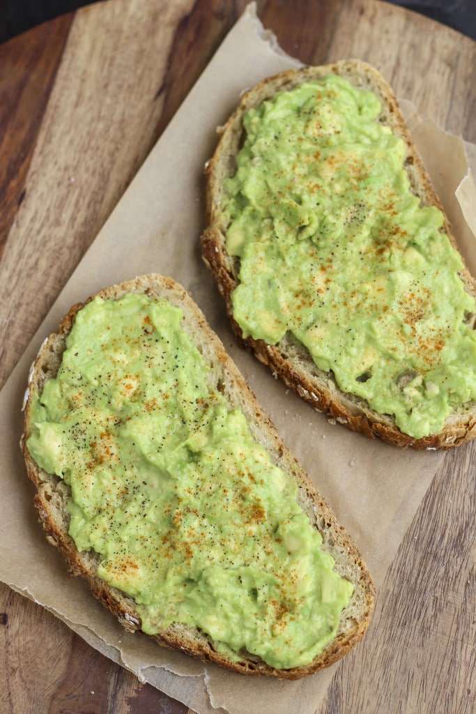 Two slices of toast with avocado on a piece of parchment paper on top of a wooden platter.