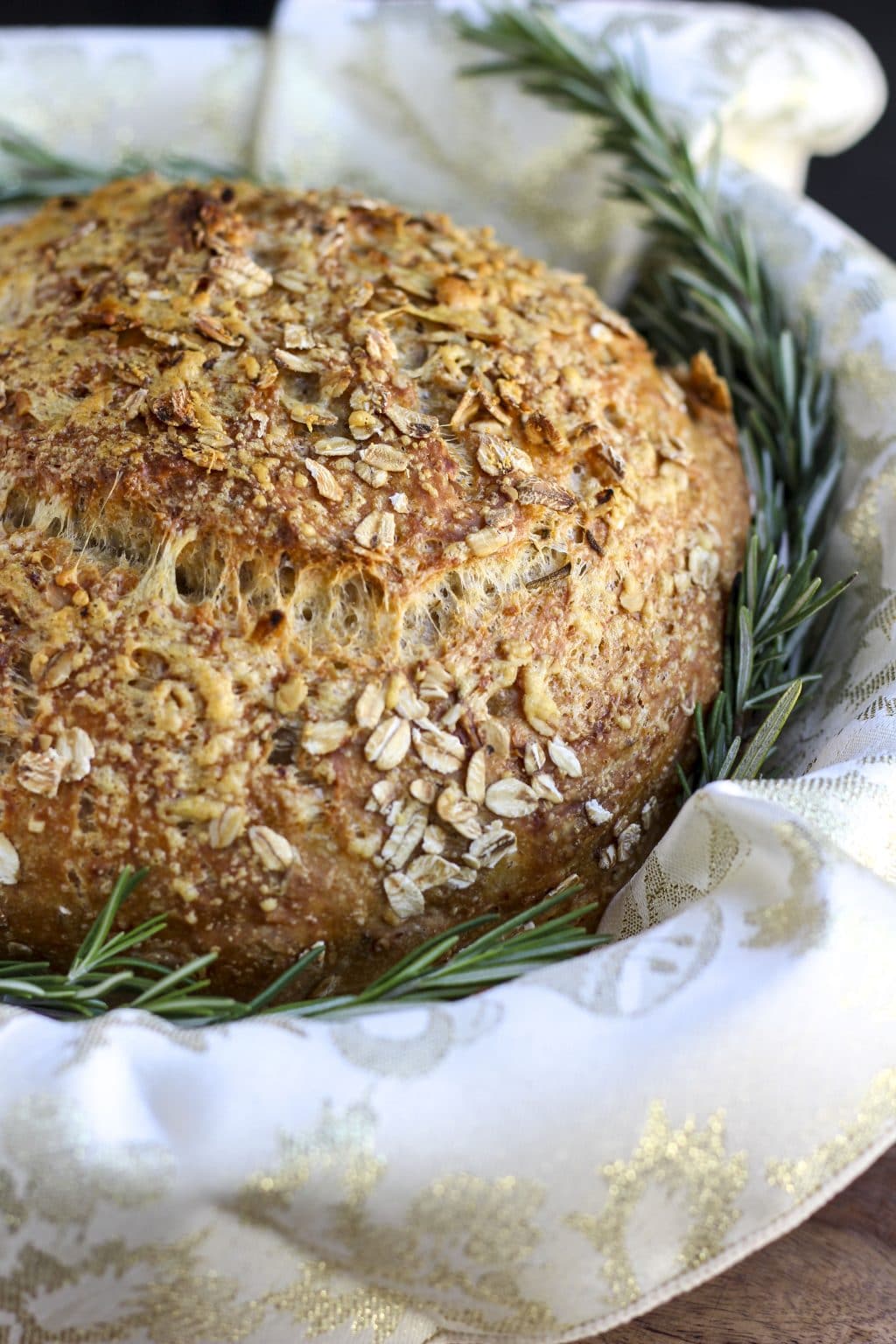 A loaf of bread in a towel lined basket with fresh rosemary springs tucked around it. 