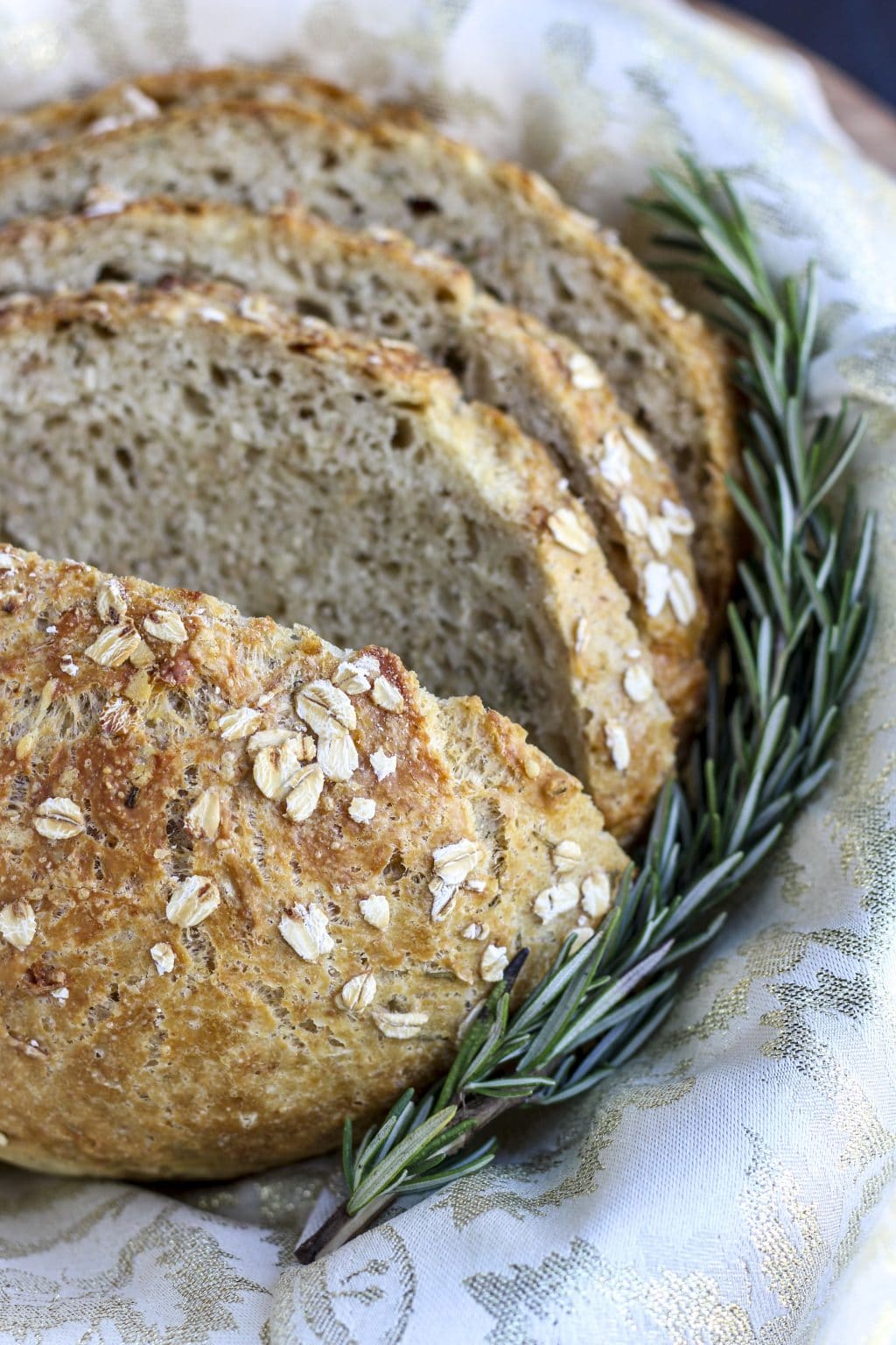 A loaf of bread cut into slices next to a sprig of rosemary in a basket. 