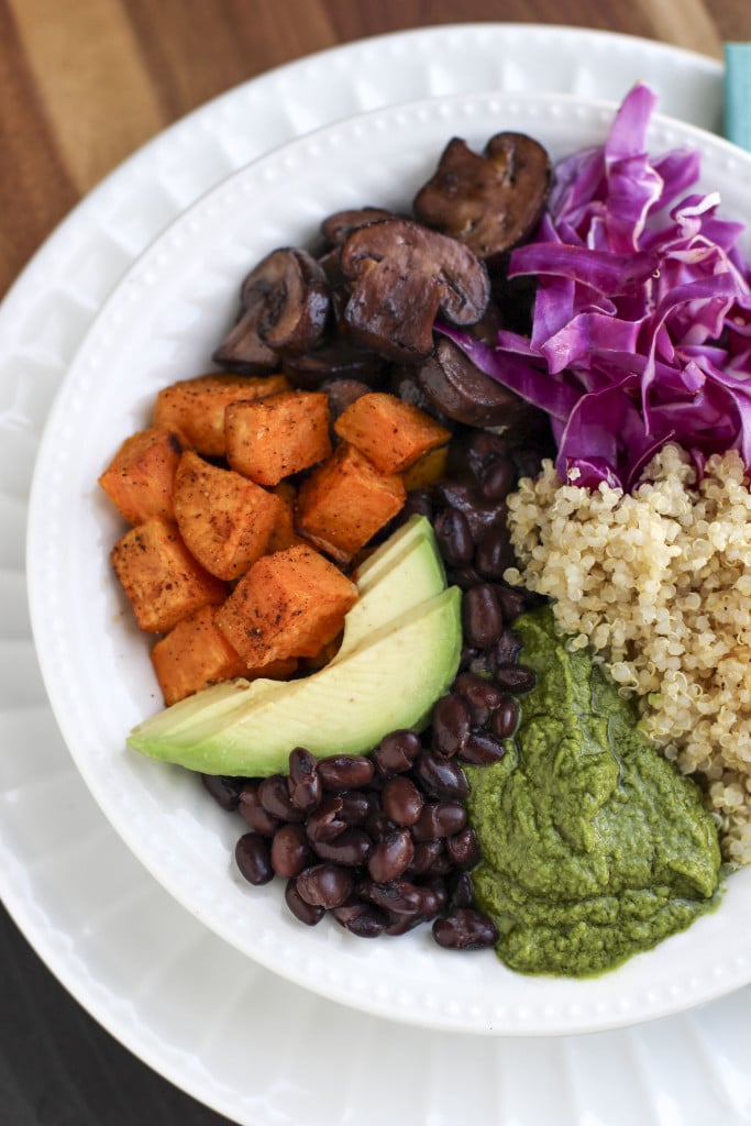 Overhead shot of a nourish bowl filled with vegetables, beans, quinoa, and green pistachio sauce on a rustic background. 