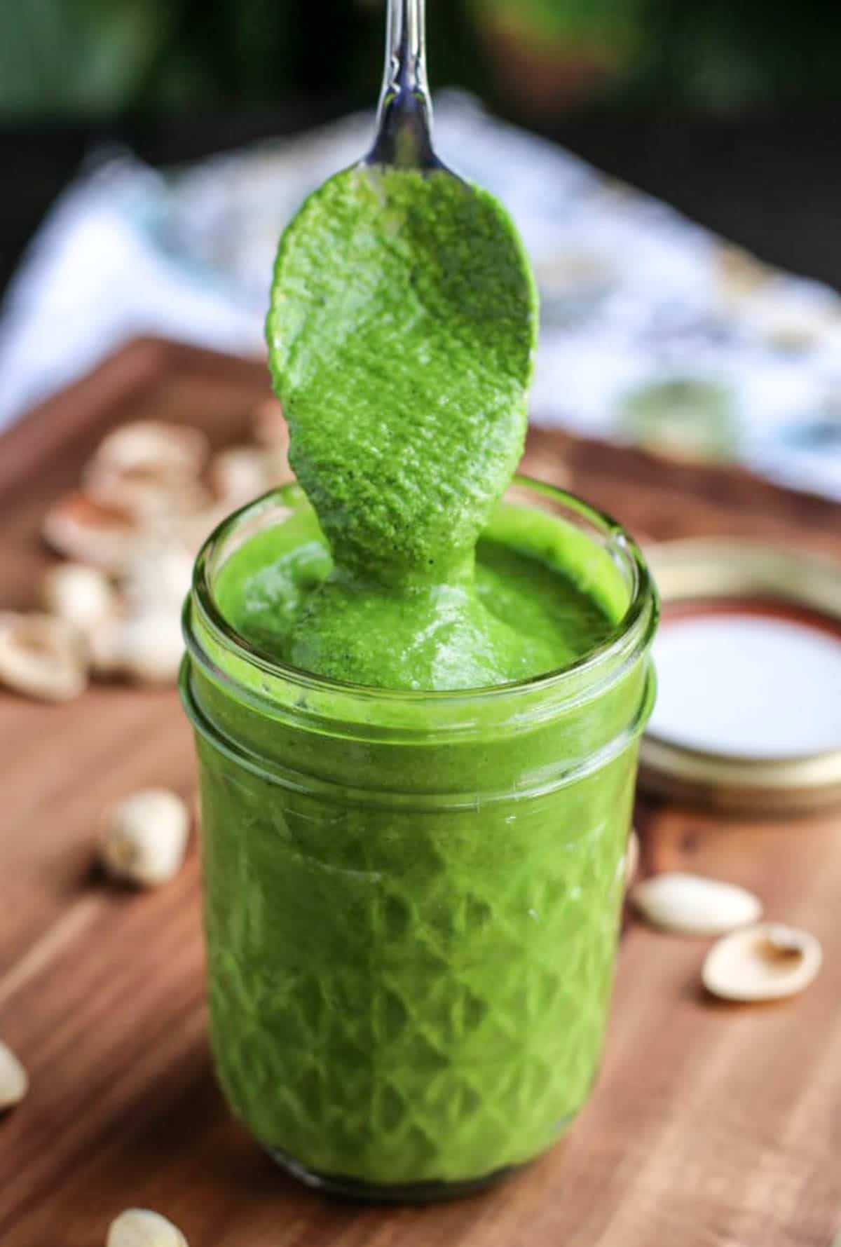 A spoon dipping into a a small mason jar filled with green pistachio sauce on a rustic background.