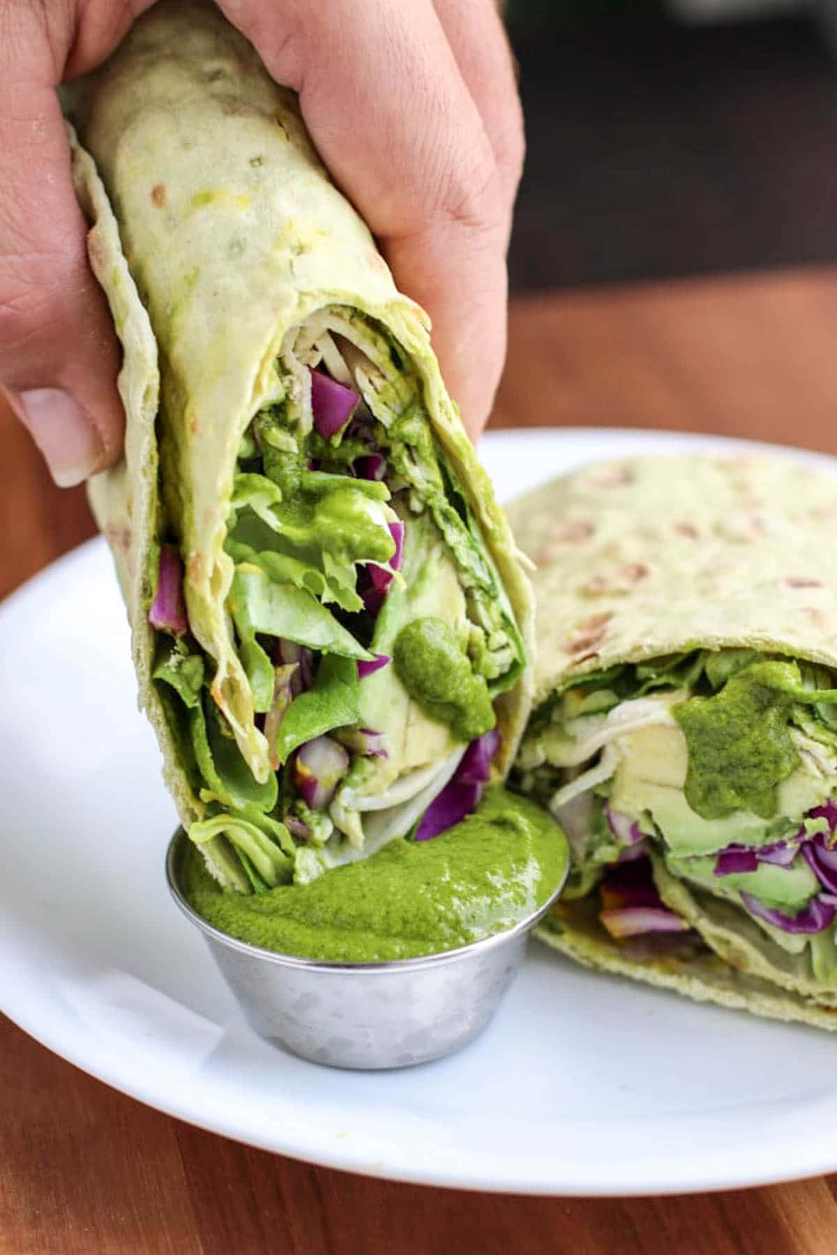 A hand dipping half a wrap into green sauce on a white plate.