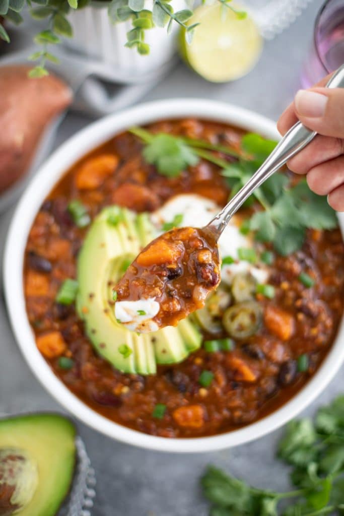 A hand holding a spoonful of chili over a bowl of food on a gray background. 