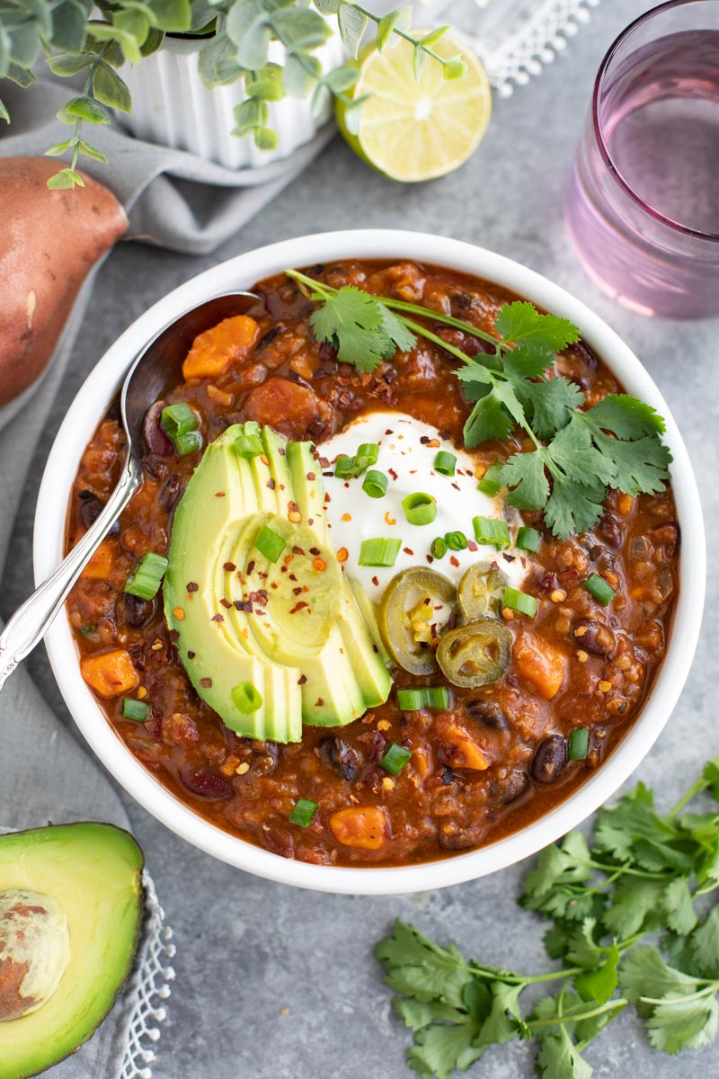 A white bowl filled with chili and a spoon next to cilantro, a cup, and a plant on a gray background.