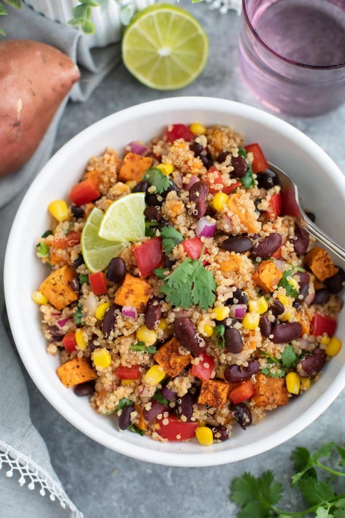 A fork in a white bowl filled with zesty quinoa salad next to half a lime and purple cup.