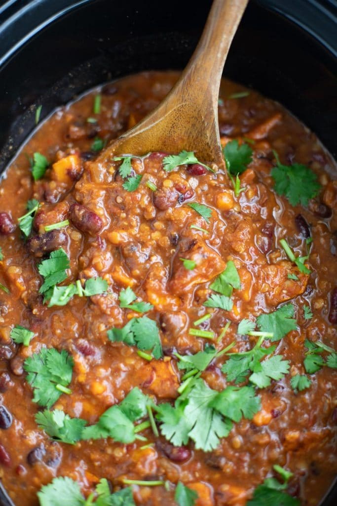 A black slow cooker filled with chili and a wooden spoon. 