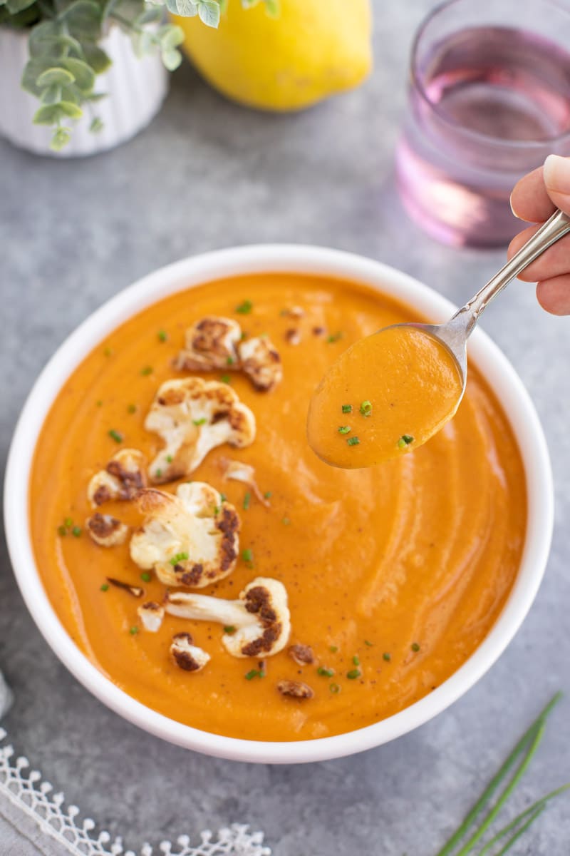 A hand dipping a spoon into a white bowl of soup on a gray background.