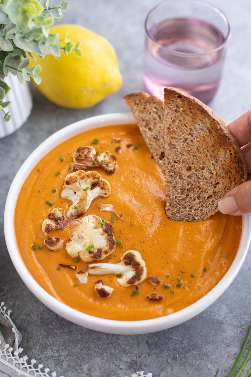 A hand dipping toast into a white bowl of soup on a gray background.