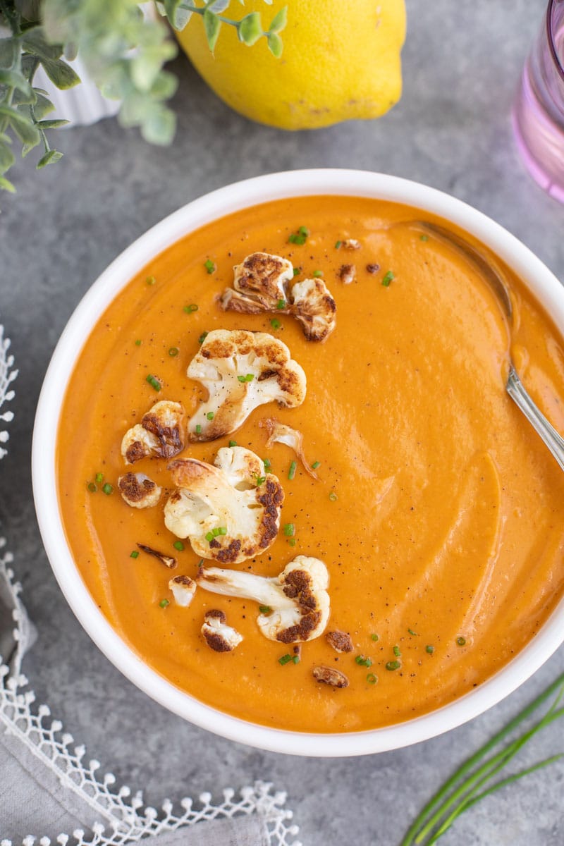 A white bowl filled with soup, a spoon, and browned cauliflower florets on a gray background.