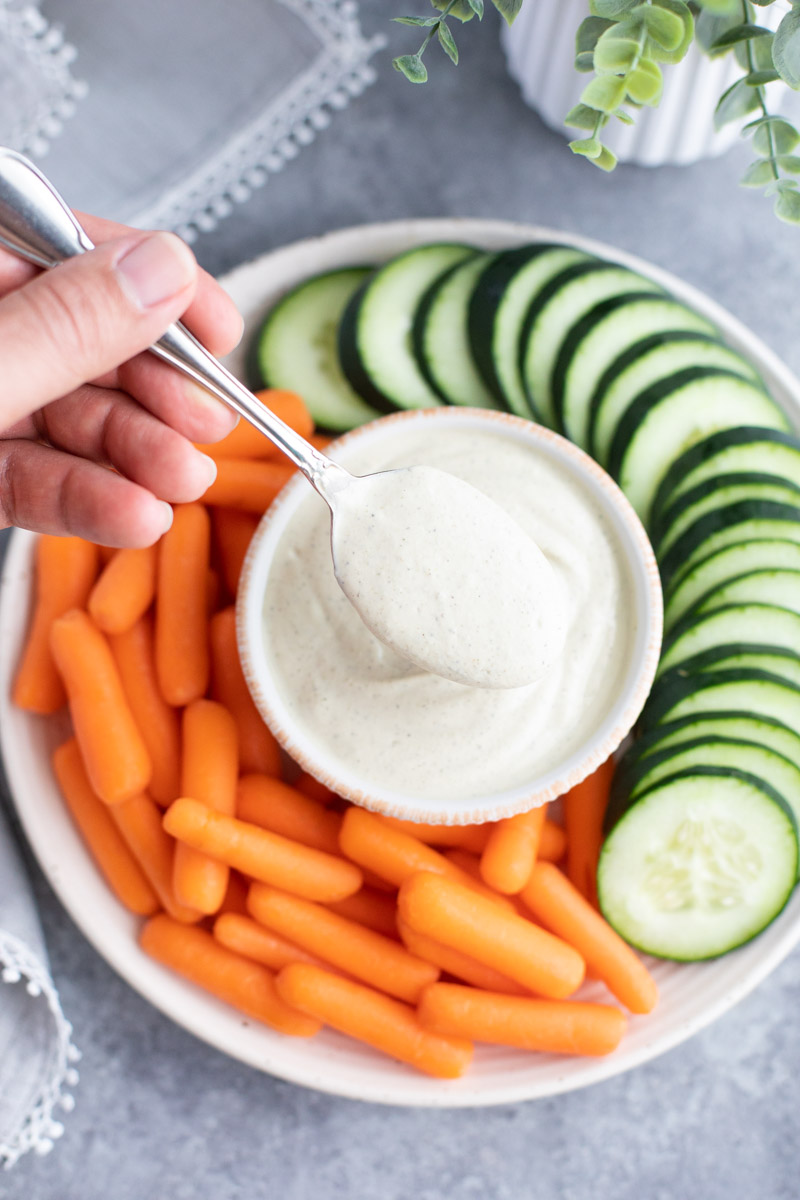 A hand holding a spoonful of dressing over a plate full of cucumbers and carrots on a gray background.