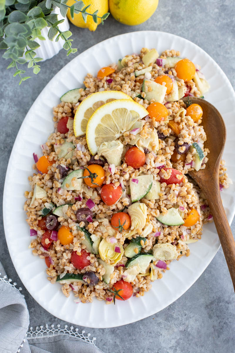 A large white platter filled with wheat berry salad next to a plant, two lemons, and a gray napkin on a gray background. 