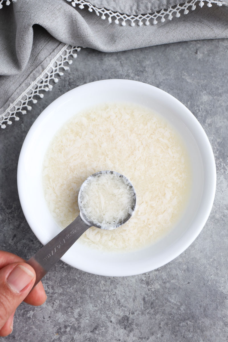 A hand holding a tablespoon of vegan buttermilk over a small bowl on a gray background. 