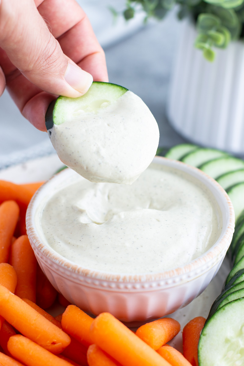 A hand dipping a cucumber slice into a bowl of vegan ranch on a plate filled with fresh vegetables. 
