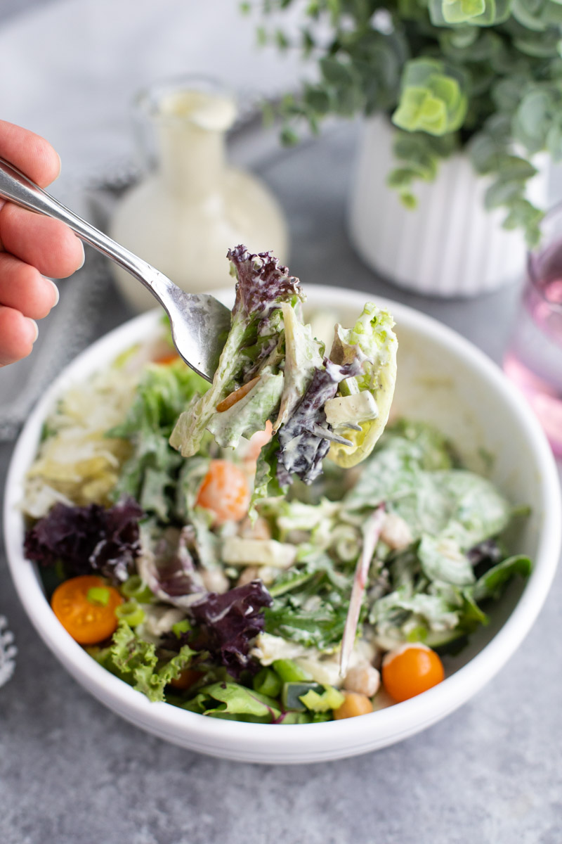 A fork full of lettuce coated in vegan ranch held over a bowl of salad on a gray background.