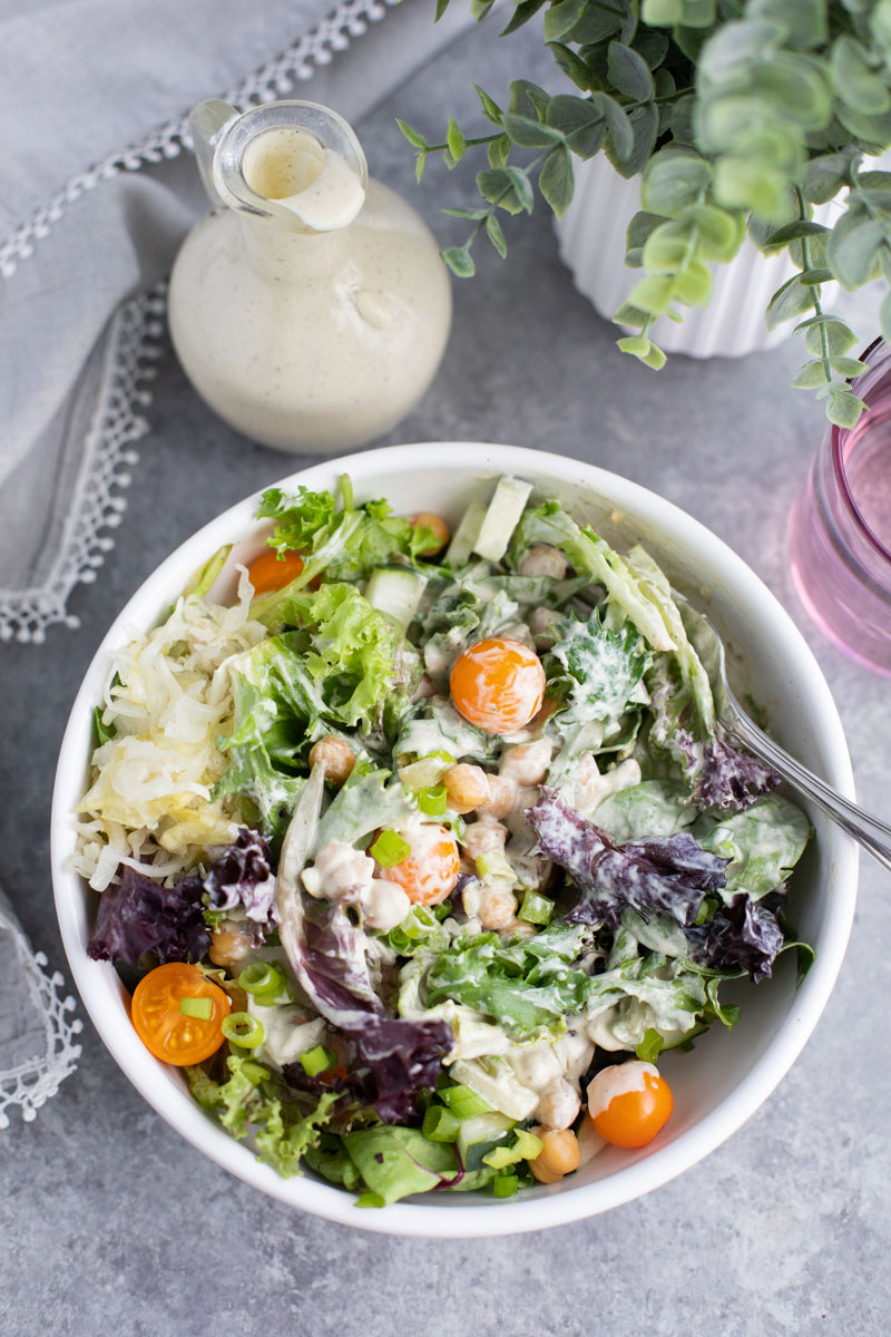 A white bowl filled with mixed greens coated in dressing next to a container of dressing on a gray background.