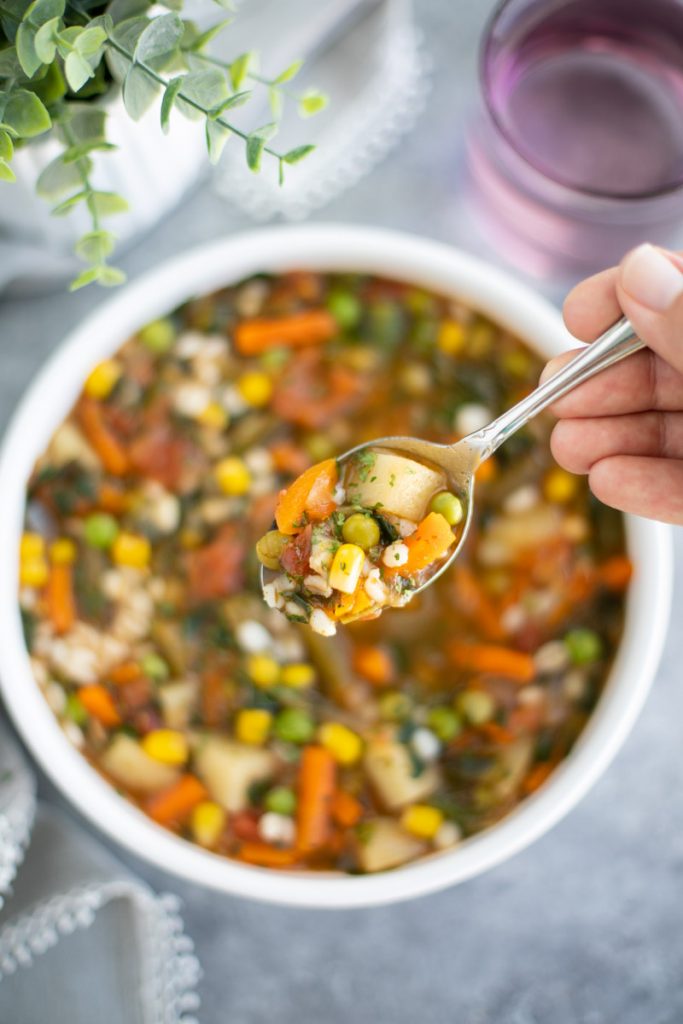 A hand holding a spoonful of soup over a filled white bowl o a gray background.