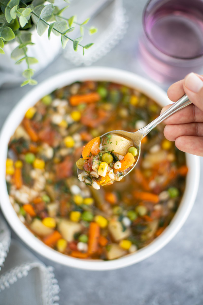 A hand holding a spoonful of soup over a filled white bowl on a gray background.