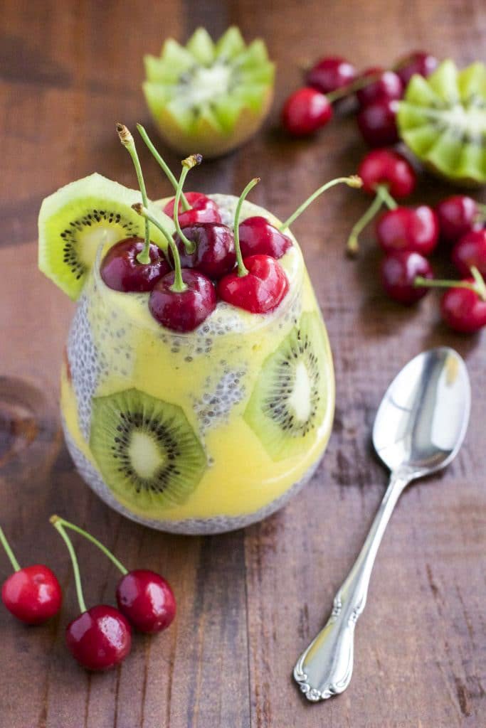 A cup filled with pudding and fruit next to fresh fruit on a wood table top.