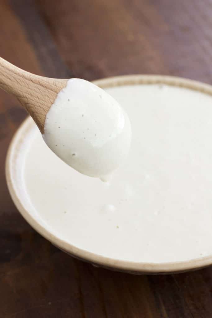 A wooden spoon being dipped into a bowl of cashew cream sauce on a rustic background. 