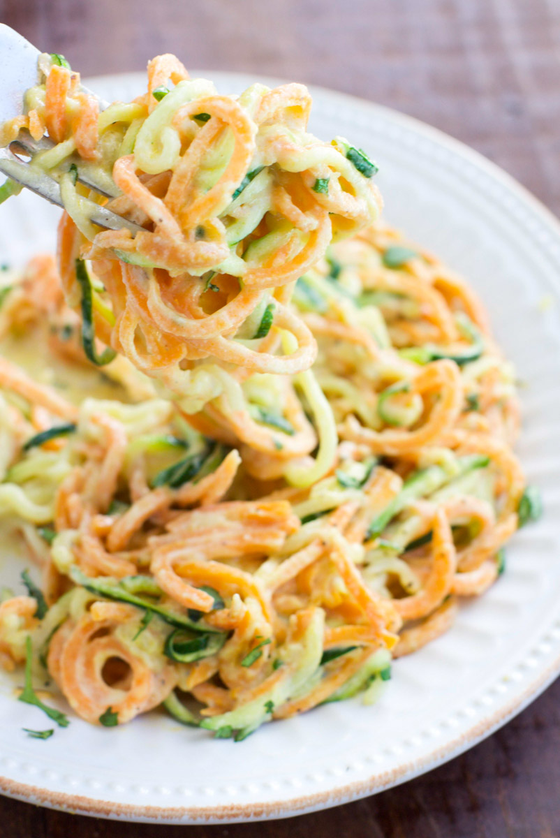 A fork holding spiraled vegetable noodles tossed in vegan white sauce over a white plate on a rustic background. 